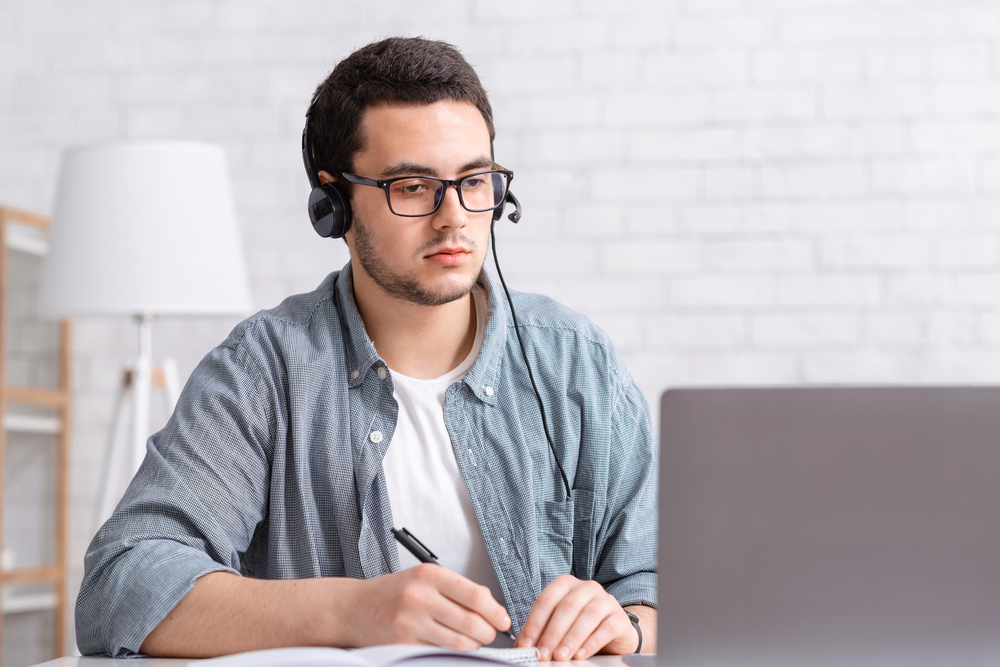 Focused guy with glasses and headphones makes notes in notebook and looks at laptop in living room interior.